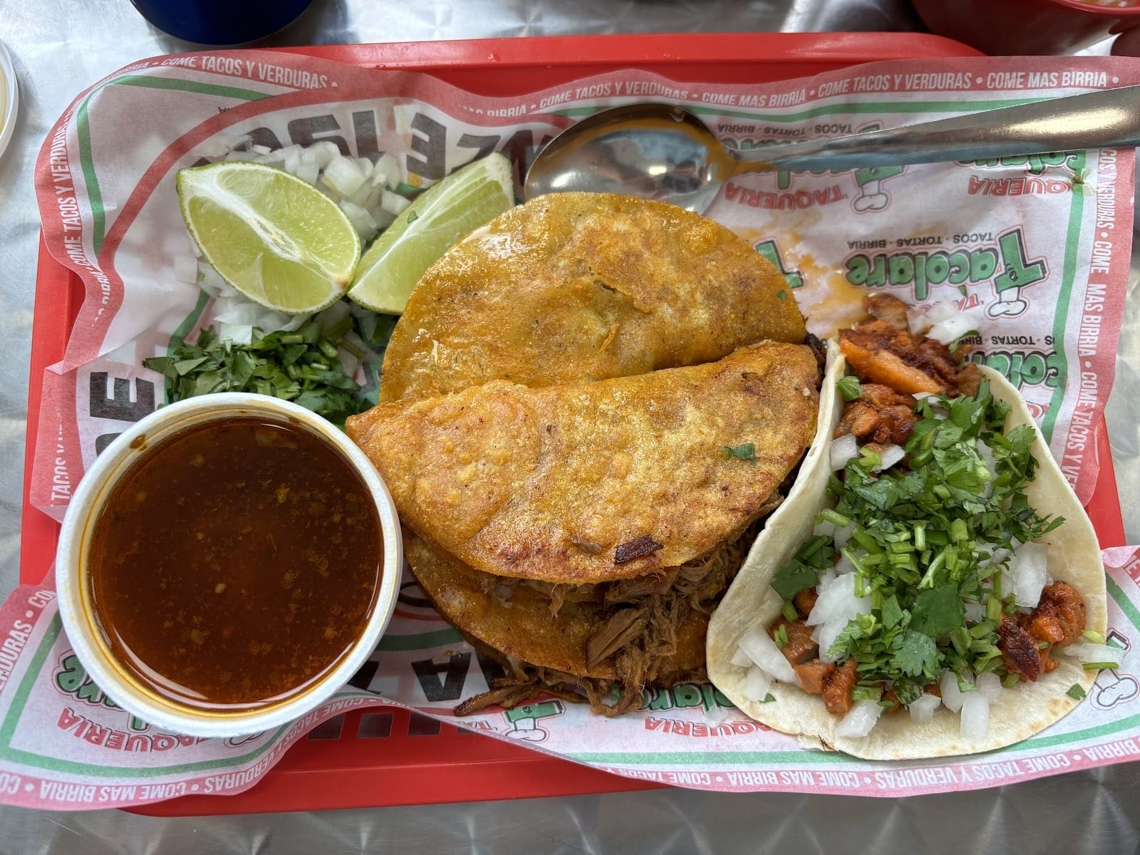 Una bandeja con comida mexicana, que incluye dos tacos de birria con una guarnición de caldo, cebolla picada, cilantro, gajos de lima y un taco pequeño. El plato se adorna con hierbas frescas y se coloca una cuchara junto a la comida.