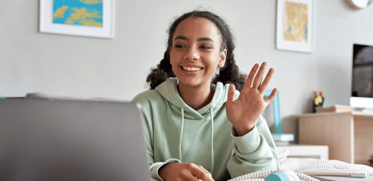 Una mujer joven está sentada en un escritorio con su ordenador portátil, sonriendo y saludando alegremente a la cámara.