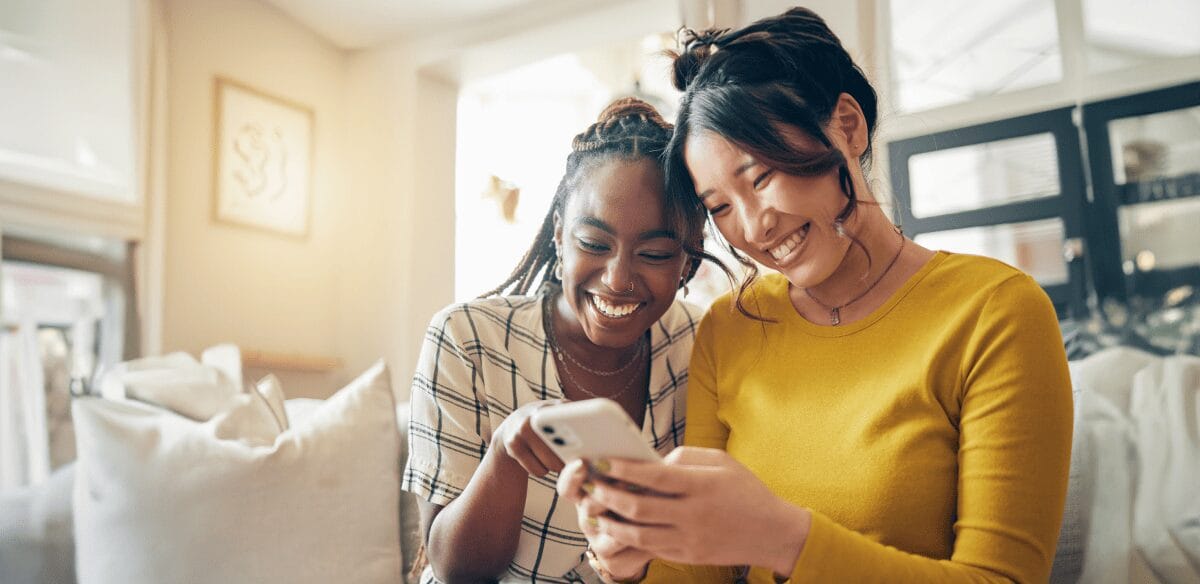 Dos mujeres sonríen y se entretienen con sus teléfonos móviles, compartiendo juntas un momento de alegría.