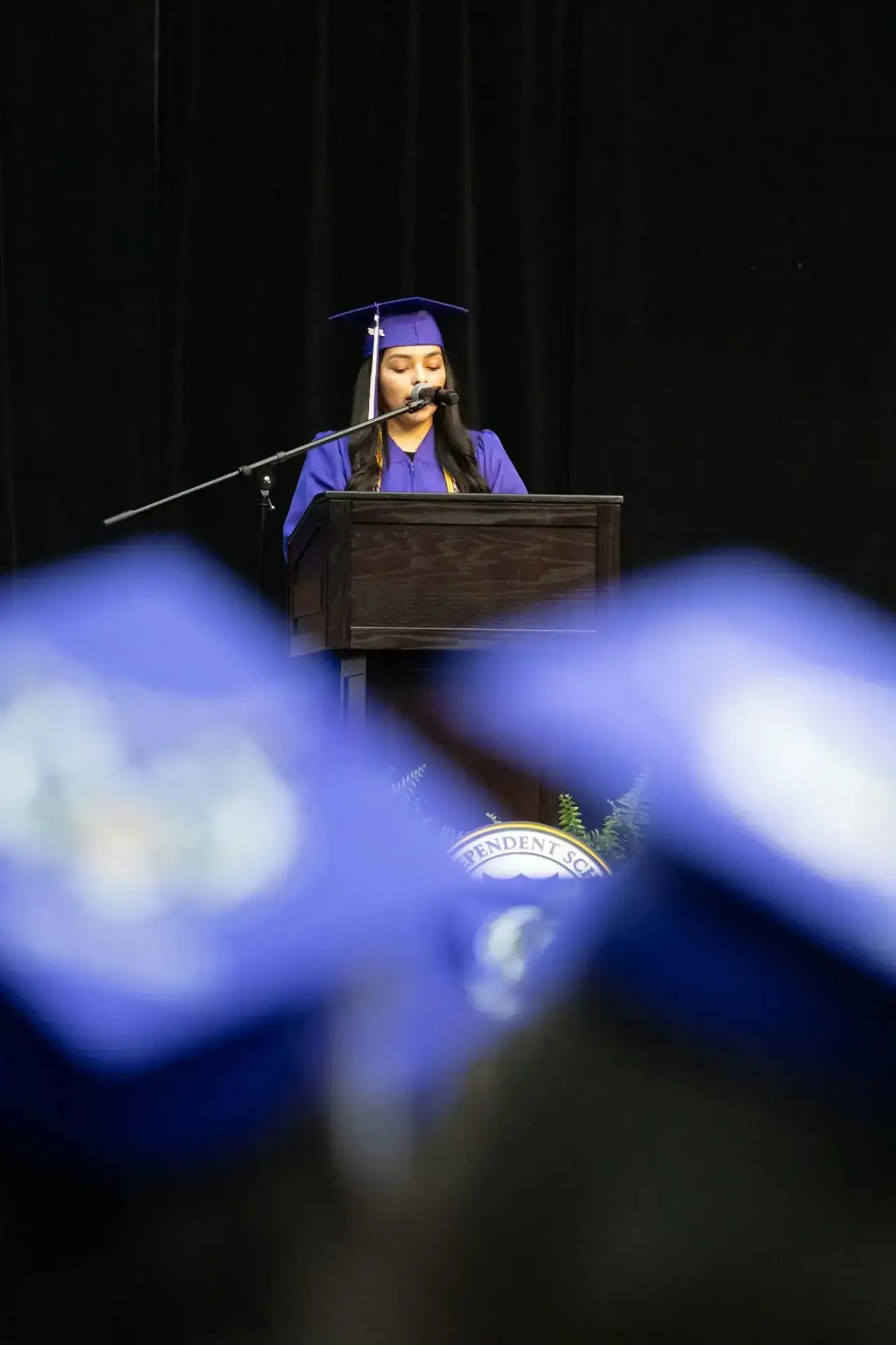 Una mujer con toga y birrete púrpura habla por un micrófono desde un podio. Las gorras azules de los graduados sentados se difuminan en primer plano. El ambiente es formal y festivo.