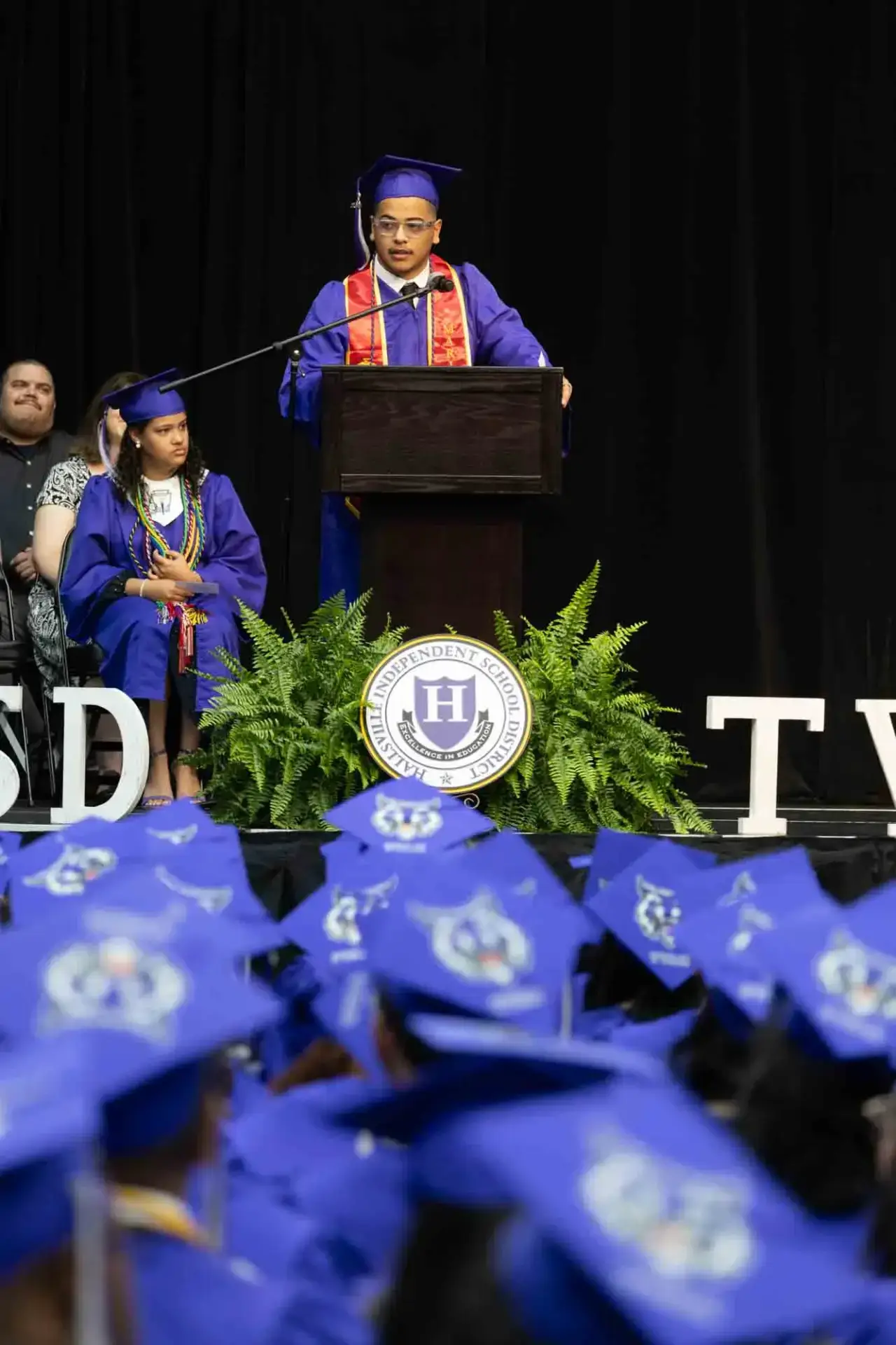 Un graduado con toga y birrete azul habla en un podio durante una ceremonia de graduación. Los graduados sentados y un emblema de la escuela son visibles, transmitiendo celebración.
