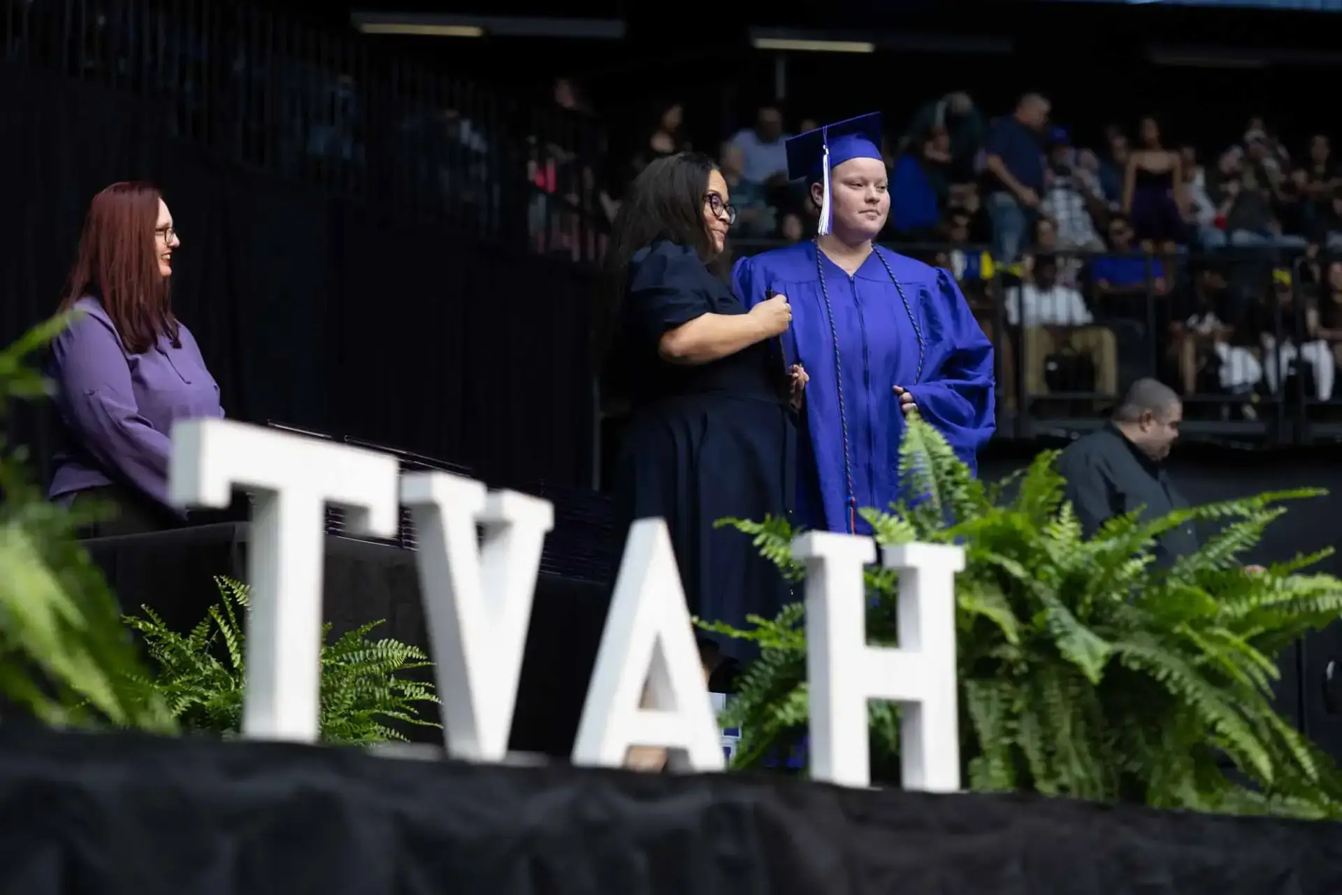 Un graduado con toga y birrete azules recibe las felicitaciones en el escenario entre frondosos helechos y grandes letras "TVAH", observado por el público en una ceremonia formal.