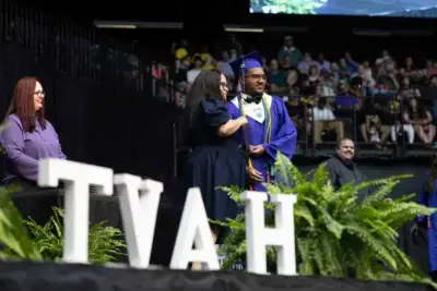 Graduado con toga y birrete recibe un diploma en el escenario de una ceremonia, sonriendo con el profesorado. Letras "TVAH" y helechos verdes adornan el primer plano.