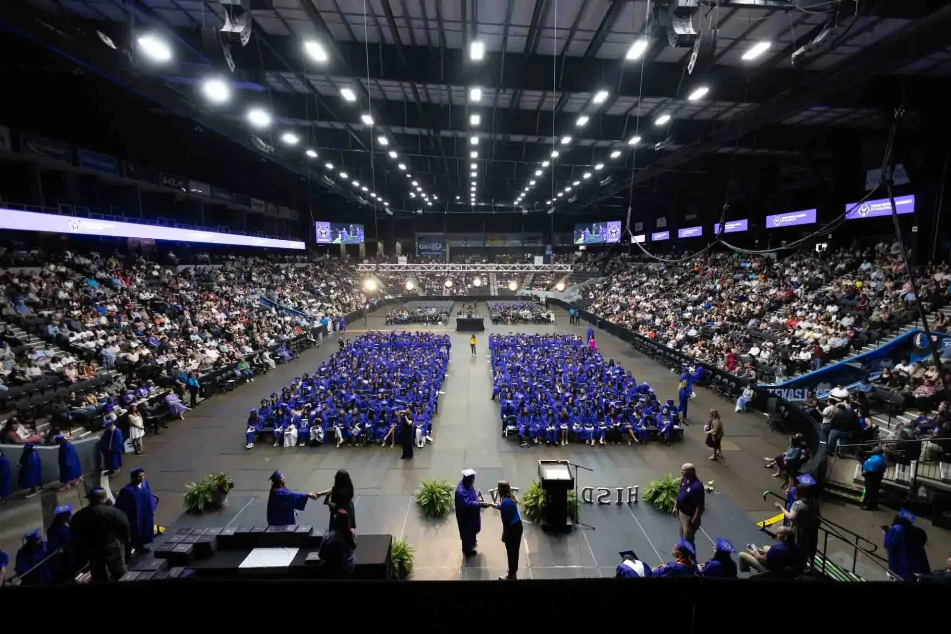 Gran angular de una ceremonia de graduación en un pabellón cubierto. Los graduados, con togas y birretes azules, están sentados en el centro, rodeados de un numeroso público. El ambiente es formal y festivo.