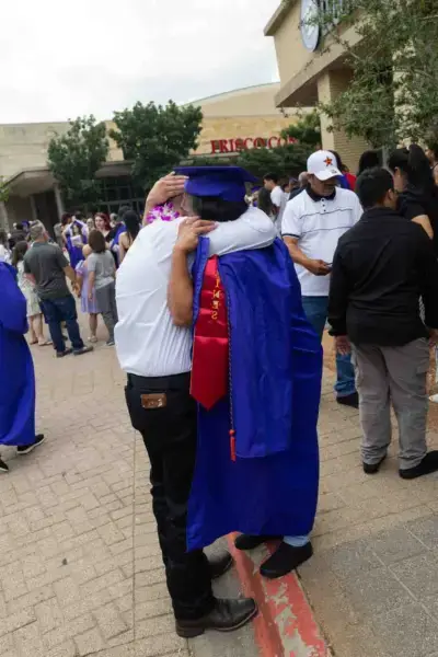 Un graduado con toga y birrete azules abraza a un adulto mayor vestido de blanco en el exterior de un local. La escena es alegre, con una multitud celebrando en el fondo.