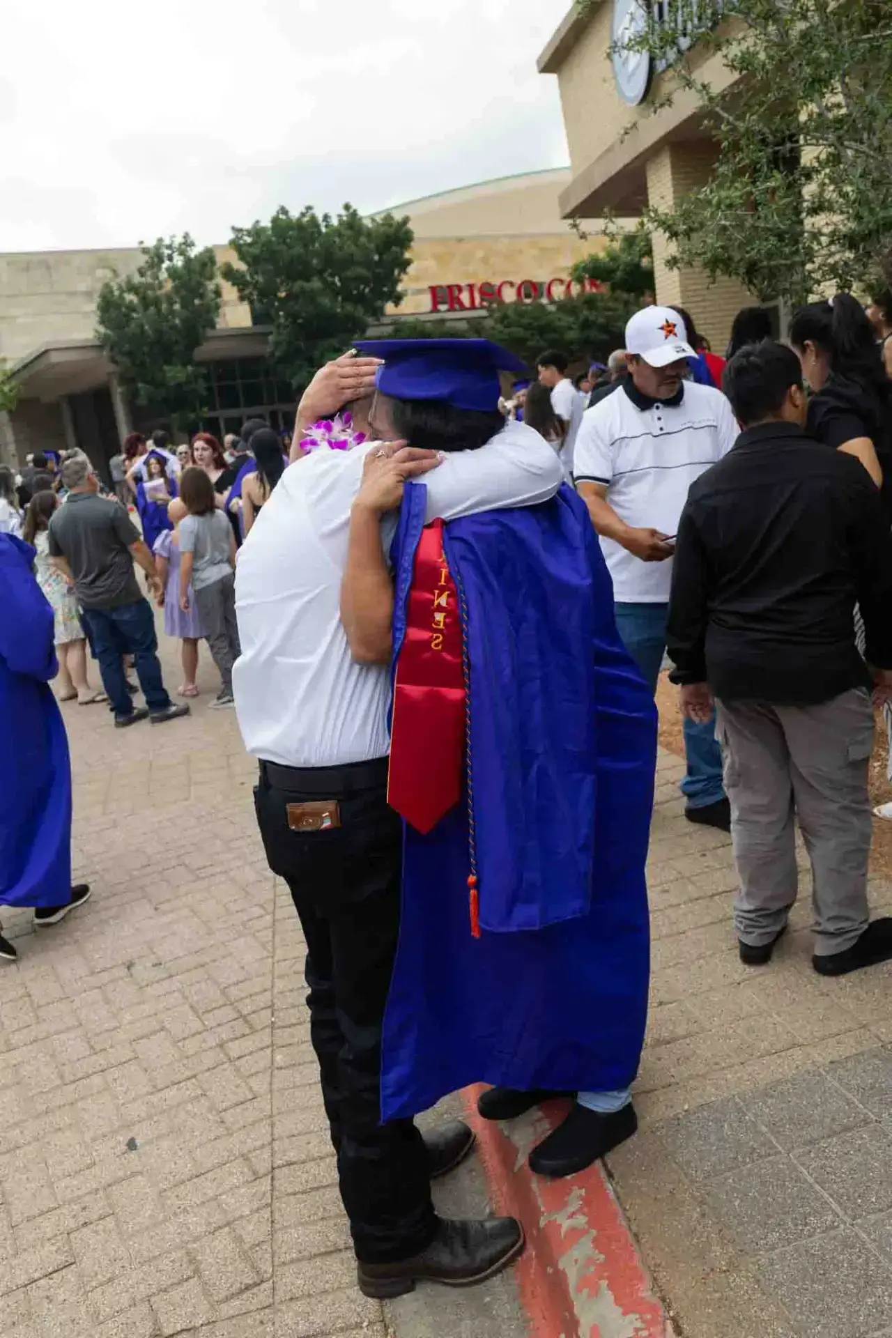 Un graduado con toga y birrete azules abraza a un adulto mayor vestido de blanco en el exterior de un local. La escena es alegre, con una multitud celebrando en el fondo.