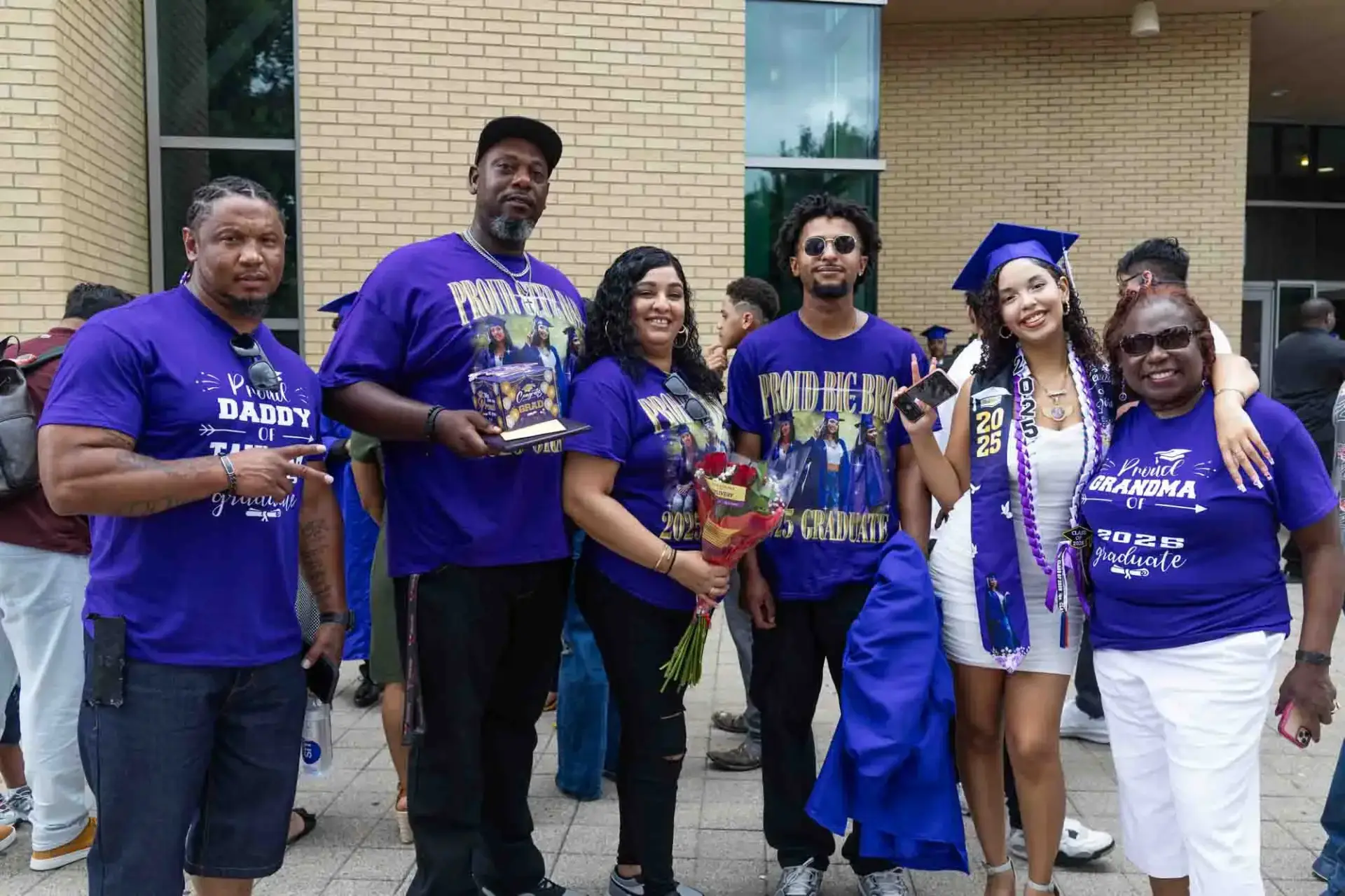 Una alegre familia celebra su graduación. Seis personas permanecen juntas en el exterior, con camisetas con mensajes de apoyo. El graduado, con toga y birrete, sonríe alegremente.
