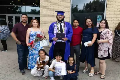 Un graduado con toga y birrete azul sostiene un diploma, rodeado de siete familiares sonrientes. Algunos sostienen flores y un osito de peluche, celebrándolo fuera.