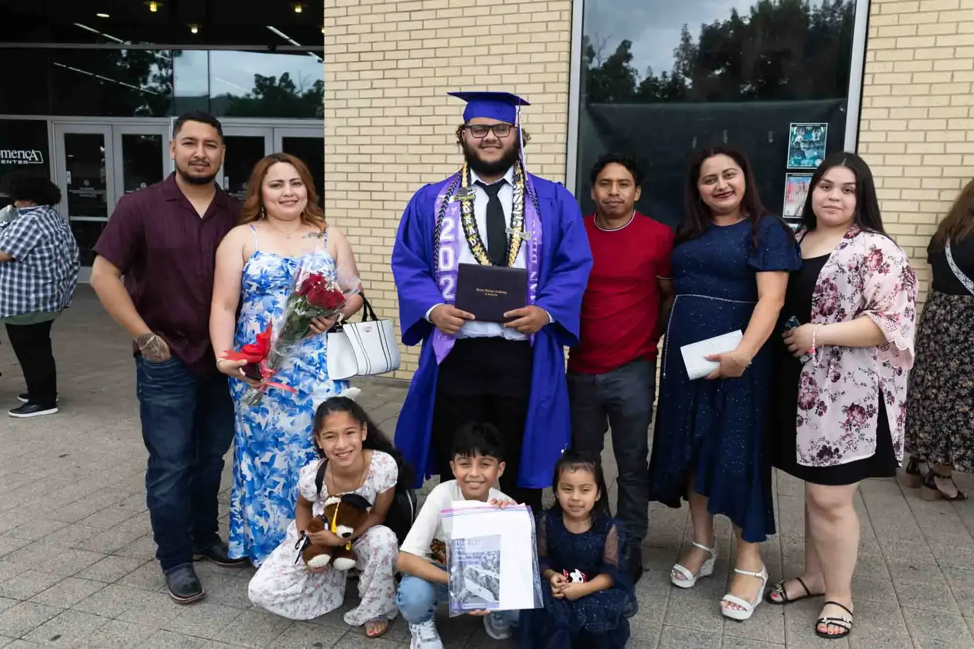Un graduado con toga y birrete azul sostiene un diploma, rodeado de siete familiares sonrientes. Algunos sostienen flores y un osito de peluche, celebrándolo fuera.