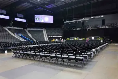 Una pista cubierta vacía con filas de sillas negras frente a un escenario. Grandes pantallas muestran logotipos y plantas verdes decoran el escenario, creando una atmósfera de expectación.