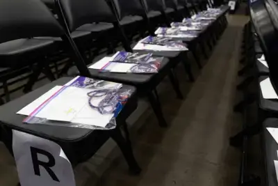 Filas de sillas negras con paquetes en cada asiento, que incluyen un folleto y un cordón de colores. Un papel con la letra "R" marca una fila. El entorno es tranquilo y organizado.