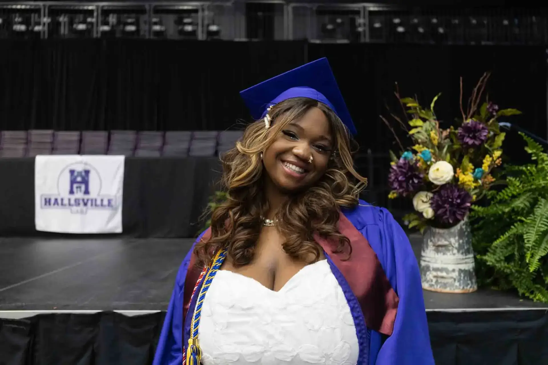 Graduada sonriente con toga y birrete azul en el escenario con la pancarta de Hallsville de fondo. Un ramo de flores de colores está cerca, transmitiendo alegría.
