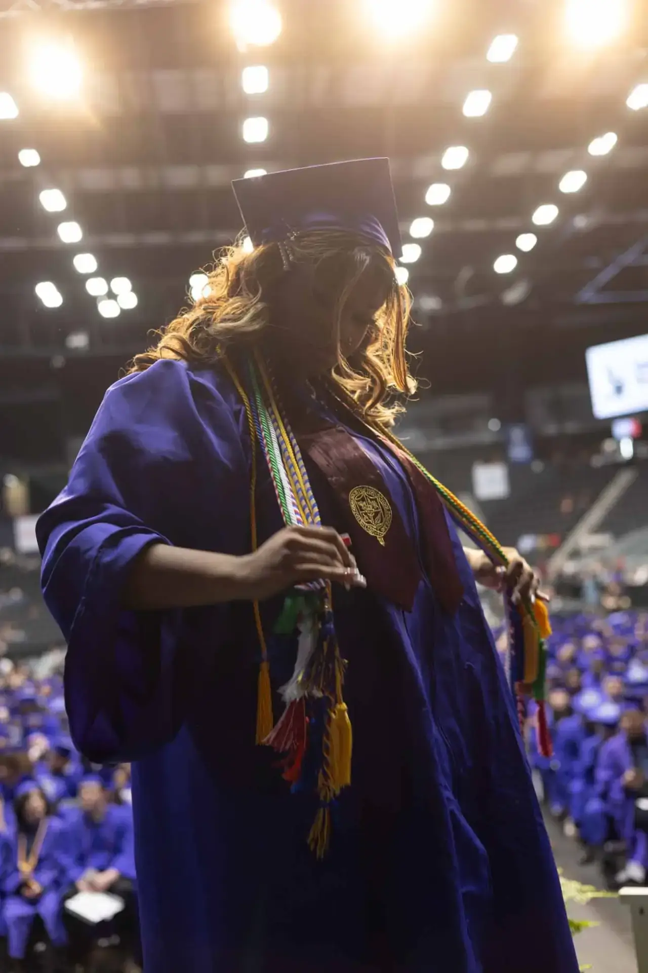 Un graduado con toga y birrete azules sostiene coloridos cordones de honor en un auditorio iluminado lleno de compañeros, transmitiendo orgullo y logro.