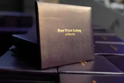 Pila de cubiertas oscuras de diplomas con texto dorado que dice "Texas Virtual Academy at Hallsville", símbolo de graduación y logro.