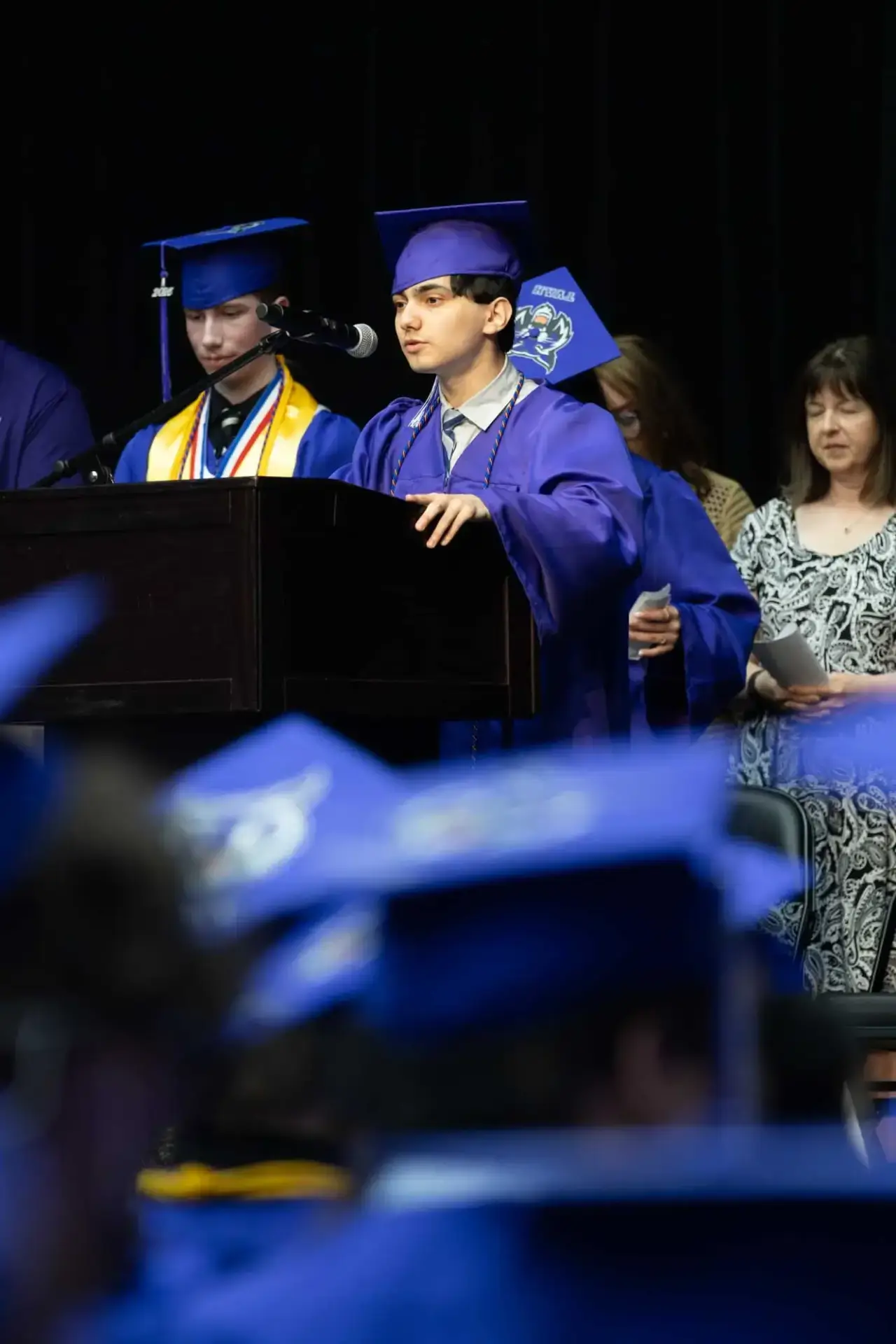 Un graduado con toga y birrete azul habla en un podio en el escenario durante una ceremonia. Un compañero se sitúa a su lado, y la gente observa al fondo.