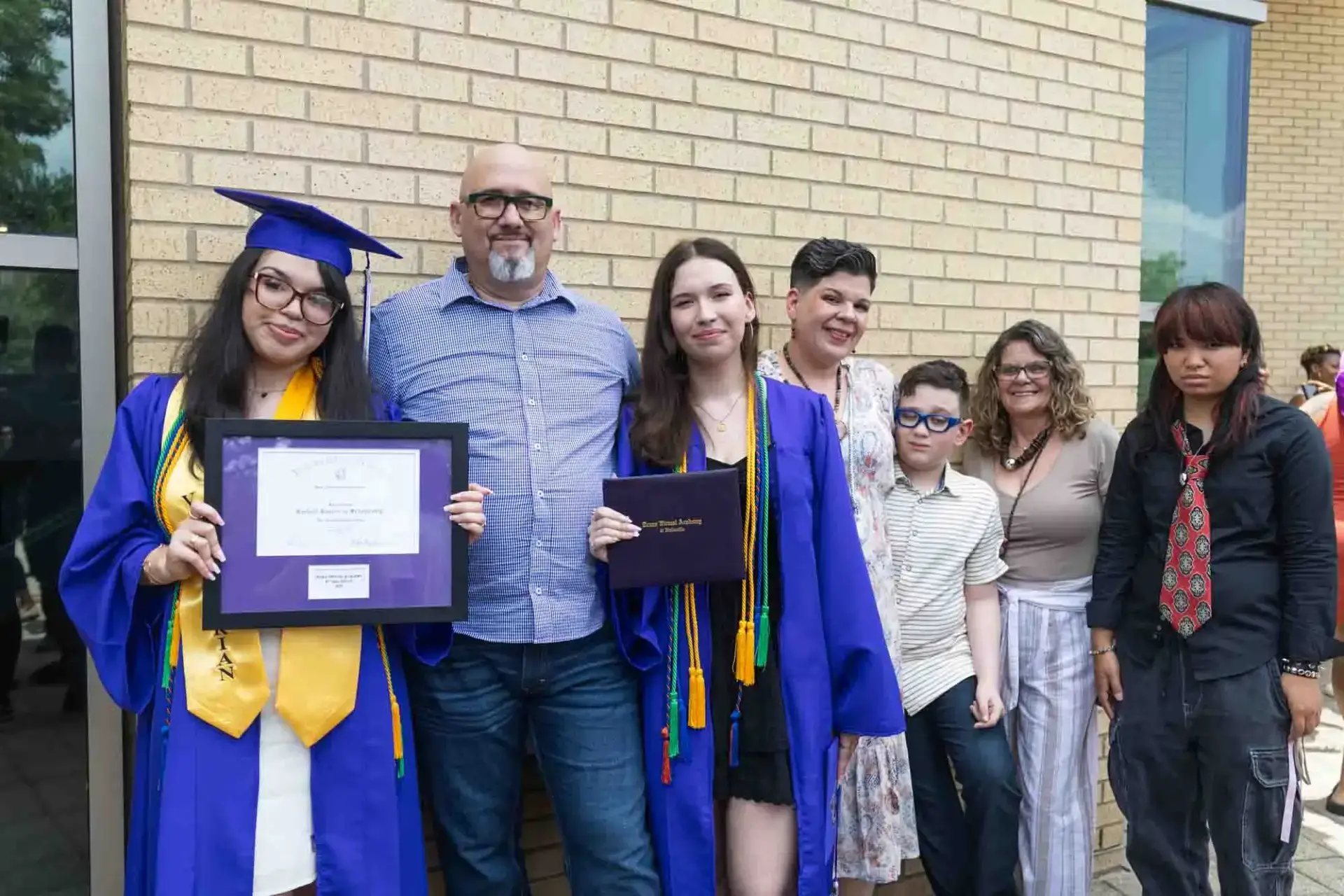 Dos graduados con toga y birrete azules sostienen sus diplomas y sonríen con orgullo. Están rodeados de familiares que les apoyan, con un muro de ladrillo como telón de fondo.