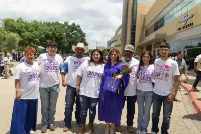 Un grupo de ocho personas posa al aire libre. Una mujer con un vestido de graduación morado sostiene flores, rodeada de familiares que la apoyan y llevan camisetas personalizadas. El ambiente es festivo.