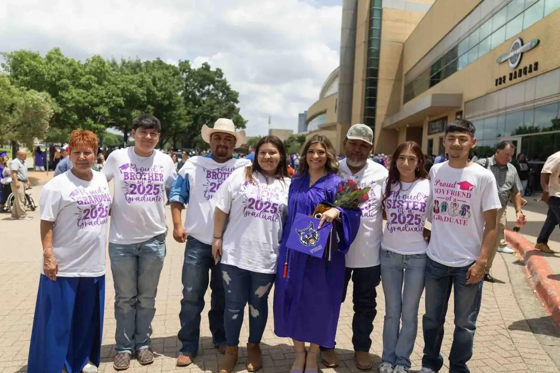 Un grupo de ocho personas posa al aire libre. Una mujer con un vestido de graduación morado sostiene flores, rodeada de familiares que la apoyan y llevan camisetas personalizadas. El ambiente es festivo.