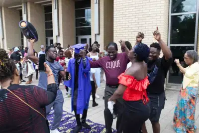 Celebración de graduación en el exterior de un edificio. Un graduado con toga y birrete azul rodeado de amigos y familiares que le aclaman, captando un momento lleno de vida.